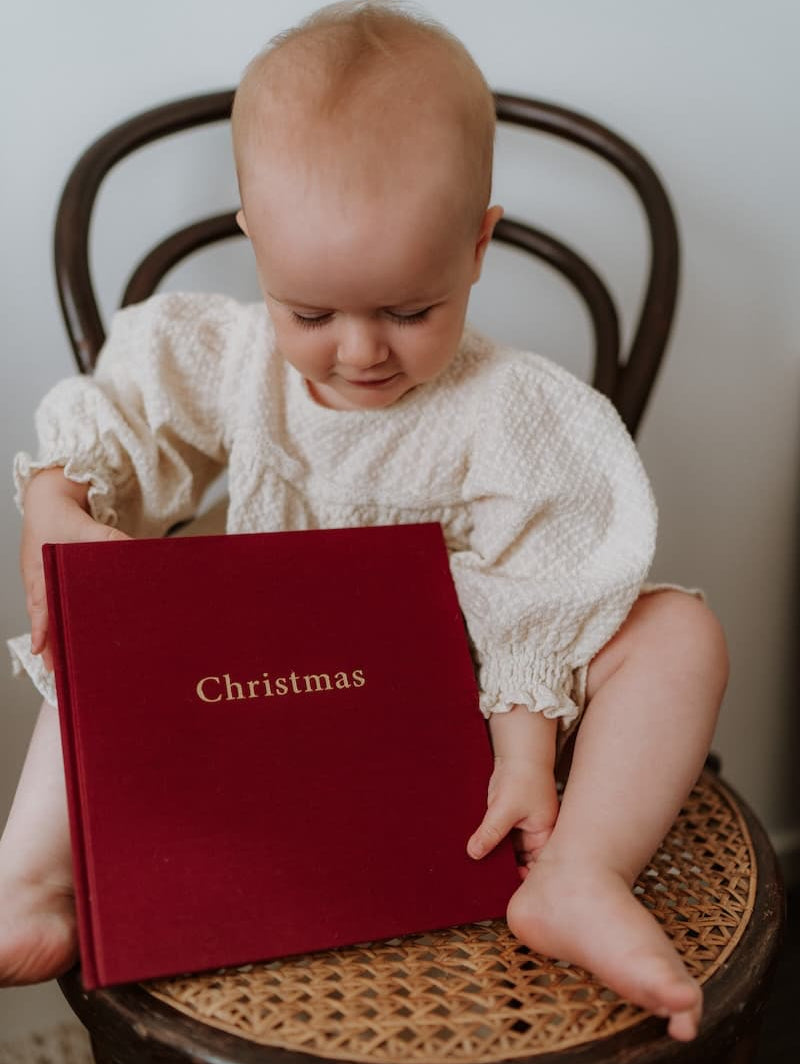 Baby holding a red family journal with 'Christmas' on it, sitting on a wicker chair.